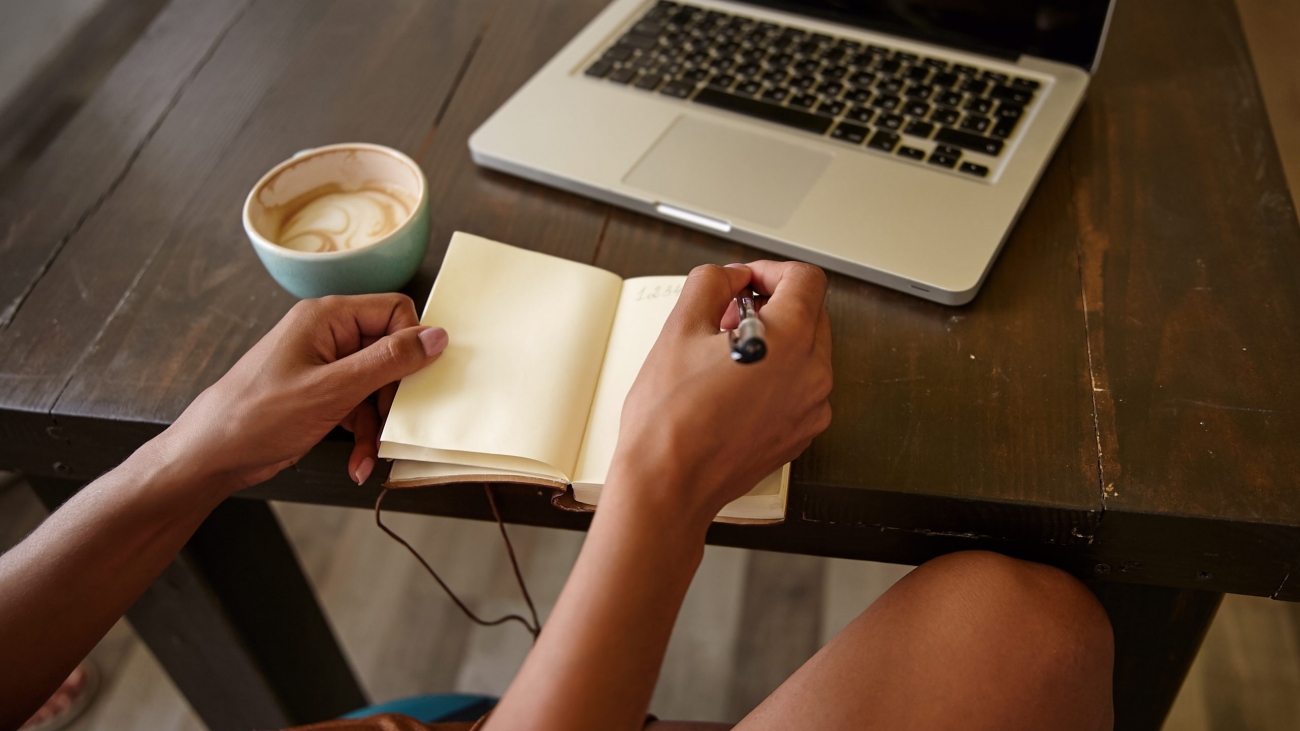 Indoor side view of female hands with pen and notebook, writing notes while having cup of coffee, using modern laptop for remote work