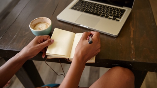 Indoor side view of female hands with pen and notebook, writing notes while having cup of coffee, using modern laptop for remote work